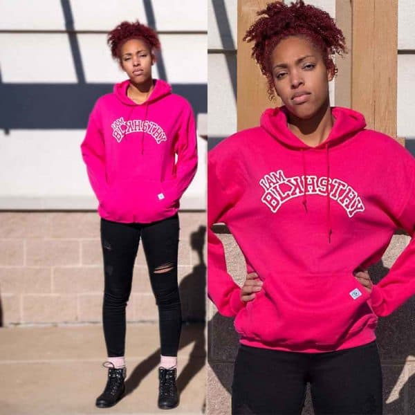 Two pictures of a College age female model standing in front of a pillar wearing pink I am black history hoodie with white letters
